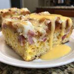 A close-up of a square slice of eggs benedict casserole topped with melted cheese and rich hollandaise sauce dripping onto a white plate.