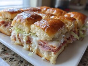 A close-up of freshly baked grinder sliders featuring melted cheese, deli meats, and shredded lettuce on shiny, glazed buns.
