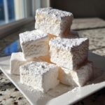 A stack of large, fluffy homemade marshmallows generously dusted with powdered sugar on a white rectangular plate.
