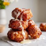 Close-up of a stack of three golden brown apple fritter bites drizzled generously with white vanilla glaze.