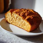 A thick, golden-brown slice of homemade soda bread resting on a white plate, catching bright sunlight.