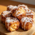 A stack of freshly baked, gooey cinnamon roll bites drizzled with thick white icing on a wooden board.