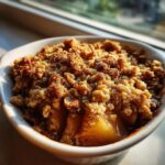 A close-up, appetizing shot of a warm individual pear crisp topped with crunchy brown crumble and nuts, sitting near a window.