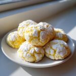 A stack of bright yellow lemon crinkle cookies heavily dusted with white powdered sugar on a white plate.