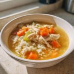 A close-up of a white bowl filled with Lemon Ginger Turmeric Chicken and Rice Soup, showing shredded chicken, rice, carrots, and celery in broth.