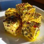 Four cubes of bright yellow marinated cheese generously coated in green herbs and red pepper flakes, resting in oil.
