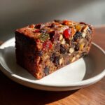 A close-up, sunlit slice of moist fruit cake packed with candied fruit and nuts on a small white plate.