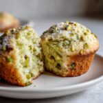 A close-up of a single pistachio muffin cut in half, revealing the moist interior filled with green pistachio pieces.
