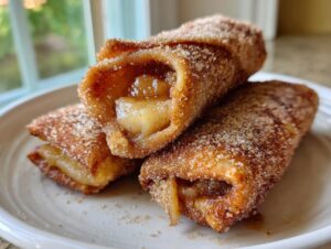 Three golden brown peach cobbler egg rolls dusted heavily with cinnamon sugar, served on a white plate.
