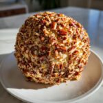 A perfectly formed cheese ball recipe coated entirely in chopped pecans, sitting on a white plate.