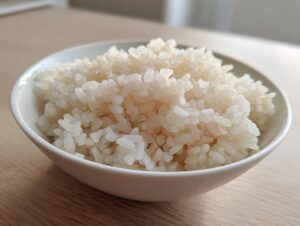 Close-up of perfectly cooked, slightly glossy sushi rice piled high in a small white bowl on a wooden surface.