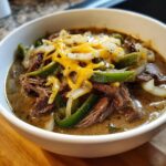 A close-up of a white bowl filled with rich philly cheese steak soup, topped with shredded beef, green peppers, onions, and melted cheese.