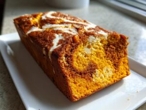 A freshly baked loaf of pumpkin cream cheese bread with a marbled cream cheese swirl on top, sitting on a white rectangular plate.