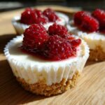 Close-up of a single raspberry topped mini cheesecake in a white paper liner, sitting on a wooden surface.