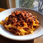 A close-up of fresh pasta topped generously with rich, slow-cooked bolognese sauce, bathed in sunlight.