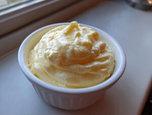 A close-up of thick, pale yellow hollandaise sauce swirled high in a small white ramekin.
