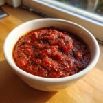 A close-up of rich, textured homemade pizza sauce seasoned with herbs, served in a white bowl.