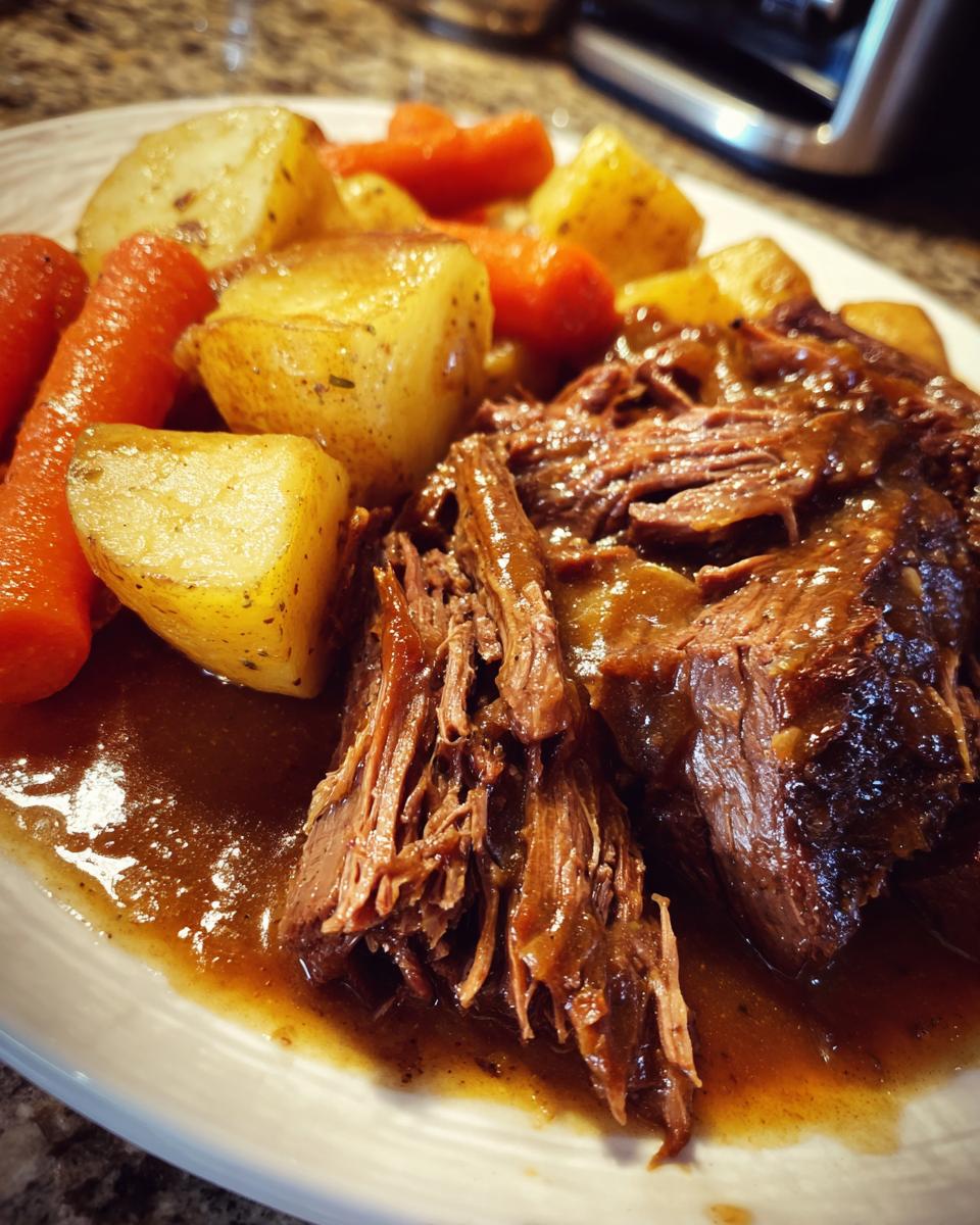Close-up of tender, shredded pot roast served with chunks of carrots and potatoes in rich brown gravy.