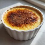 Close-up of a silky egg custard in a white fluted ramekin, featuring a beautifully torched, caramelized top dusted with cocoa powder.