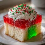 A close-up of a slice of christmas poke cake featuring white cake, red and green jello layers, whipped cream, and holiday sprinkles.