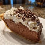 A close-up of a rich slice of french silk pie featuring a thick chocolate mousse filling, topped with whipped cream and chocolate shavings.