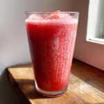 A tall glass filled with a vibrant, icy watermelon smoothie, sitting on a sunlit wooden surface.