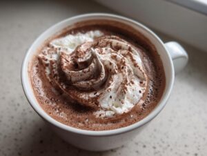 A close-up of a mug filled with rich, frothy whipped hot chocolate topped with a swirl of cream and dusted with cocoa powder.
