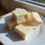 Several squares of creamy, pale white chocolate fudge stacked on a white plate near a sunny window.