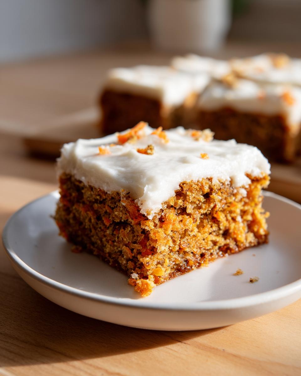 A close-up of a moist slice of carrot cake bars topped with thick cream cheese frosting and grated carrot.