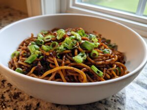 A white bowl filled with glossy, dark brown garlic noodles, topped generously with bright green sliced scallions.