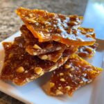 A stack of broken, shiny pieces of homemade peanut brittle recipe on a white plate.