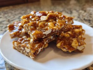 A stack of freshly broken pieces of shiny, golden-brown peanut brittle recipe candy on a white plate.