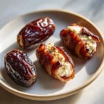 Two bacon wrapped stuffed dates filled with creamy cheese sit next to two plain dates on a small plate.