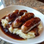 A close-up of bangers and mash featuring four browned sausages topped with onion gravy, served over creamy mashed potatoes.