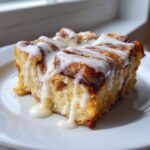 A close-up of a serving of cinnamon roll french toast dripping with thick white glaze on a white plate.