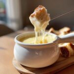 A piece of crusty bread being dipped into a white pot of hot, stretchy cheese fondue recipe.