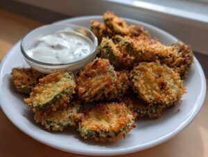 A plate piled high with golden brown, crunchy Fried Pickles next to a small bowl of creamy white dipping sauce.