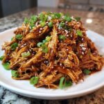 A mound of glossy, shredded crockpot teriyaki chicken topped with green onions and sesame seeds on a white plate.