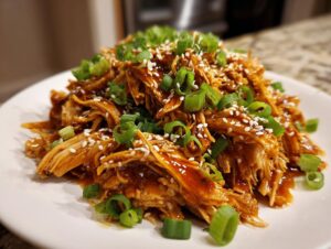 Close-up of shredded crockpot teriyaki chicken coated in sauce, topped with green onions and sesame seeds.