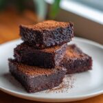 A stack of four fudgy date brownies dusted generously with cocoa powder on a white plate.