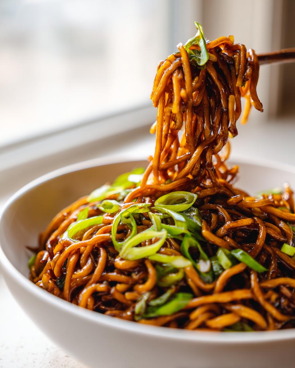 Chopsticks lifting a generous portion of glossy, dark brown garlic noodles topped with bright green scallions from a white bowl.
