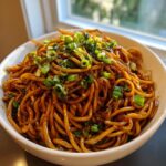A close-up of a white bowl filled with glossy, dark sauced garlic noodles, garnished generously with chopped green onions.