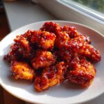 Close-up of crispy, glazed gochujang chicken pieces piled high on a white plate near a window.