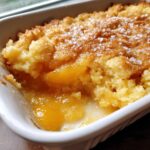 Close-up of a warm, golden peach cobbler in a white dish, showing bubbling fruit filling and a crumbly topping sprinkled with coarse sugar.