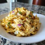 A mound of creamy, yellow-tinted honey mustard chicken salad with visible chunks of shredded chicken, red onion, and celery in a white bowl.