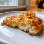 Close-up of a flaky, golden-brown Parmesan crusted tilapia fillet on a white plate.