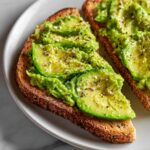 Close-up of two slices of avocado toast topped with fresh avocado slices and coarse black pepper.