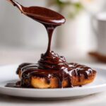 A copper spoon pours thick, glossy chocolate syrup over a small baked dessert on a white plate.