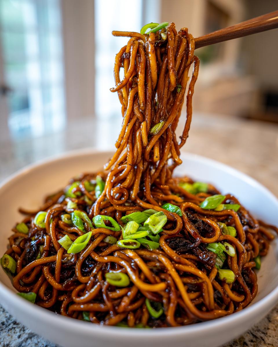 A close-up shot of glossy, dark sauce-coated garlic noodles being lifted from a white bowl with chopsticks, topped with fresh green onions.