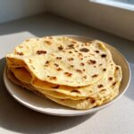 A stack of freshly cooked, slightly charred corn tortillas resting on a white plate in natural sunlight.